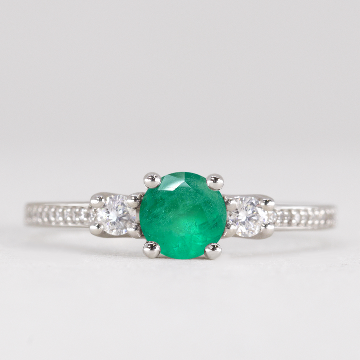 Round cut emerald and diamond trilogy engagement ring with white diamond pave band